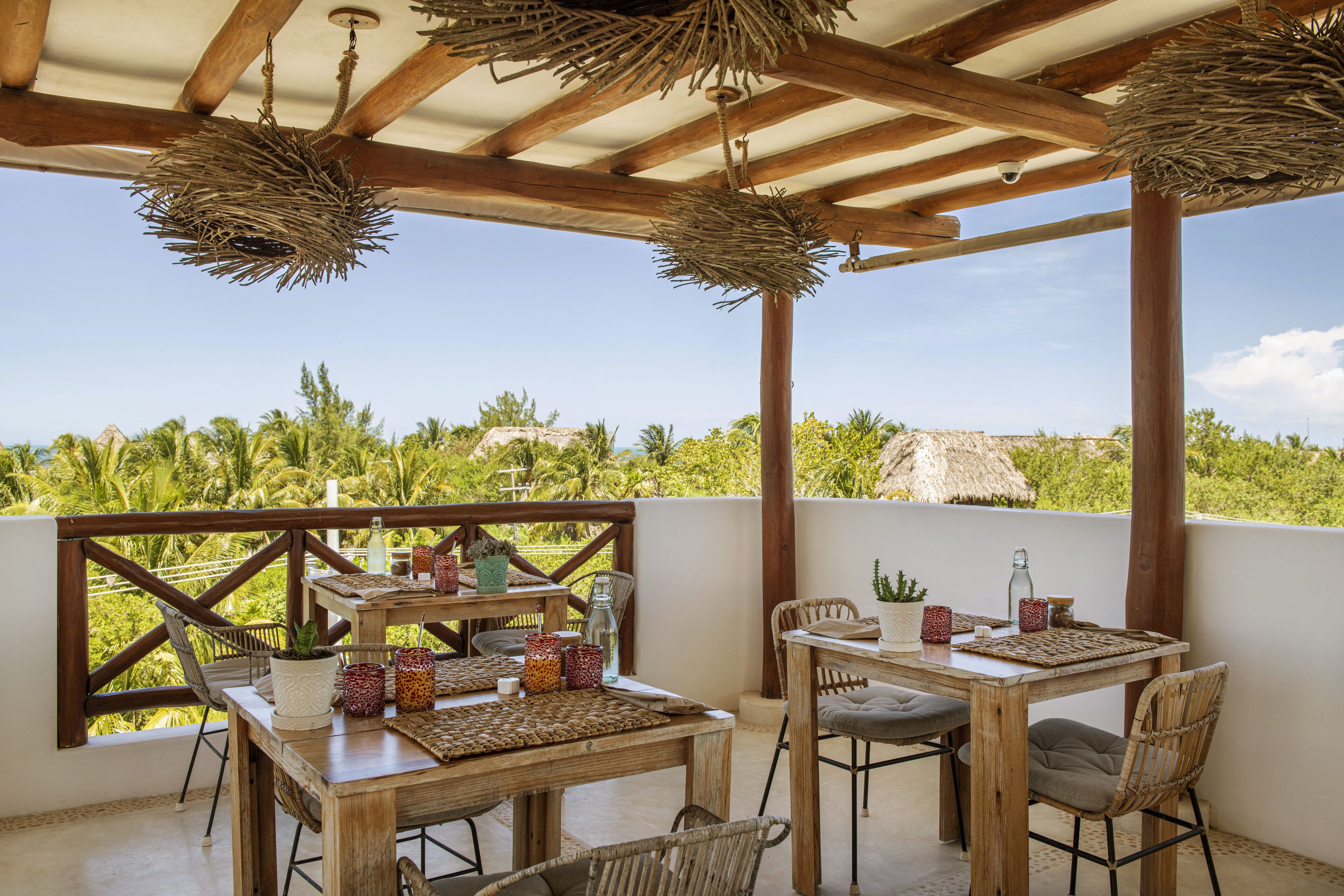 An outdoor patio with wooden tables and chairs, surrounded by lush greenery and a clear sky.