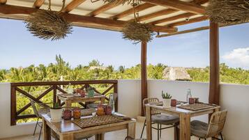 An outdoor patio with wooden tables and chairs, surrounded by lush greenery and a clear sky.