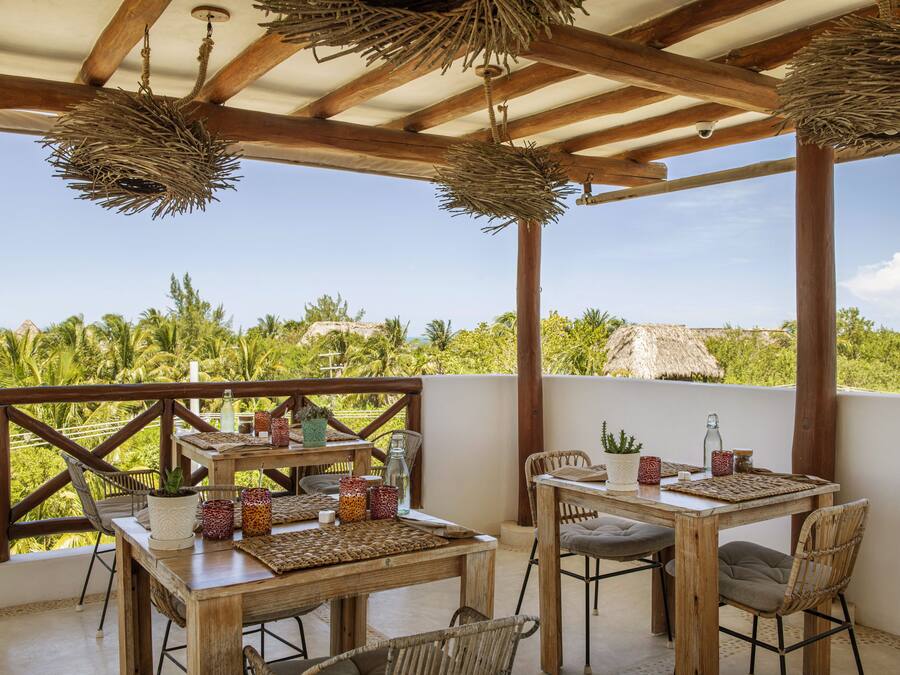 An outdoor patio with wooden tables and chairs, surrounded by lush greenery and a clear sky.