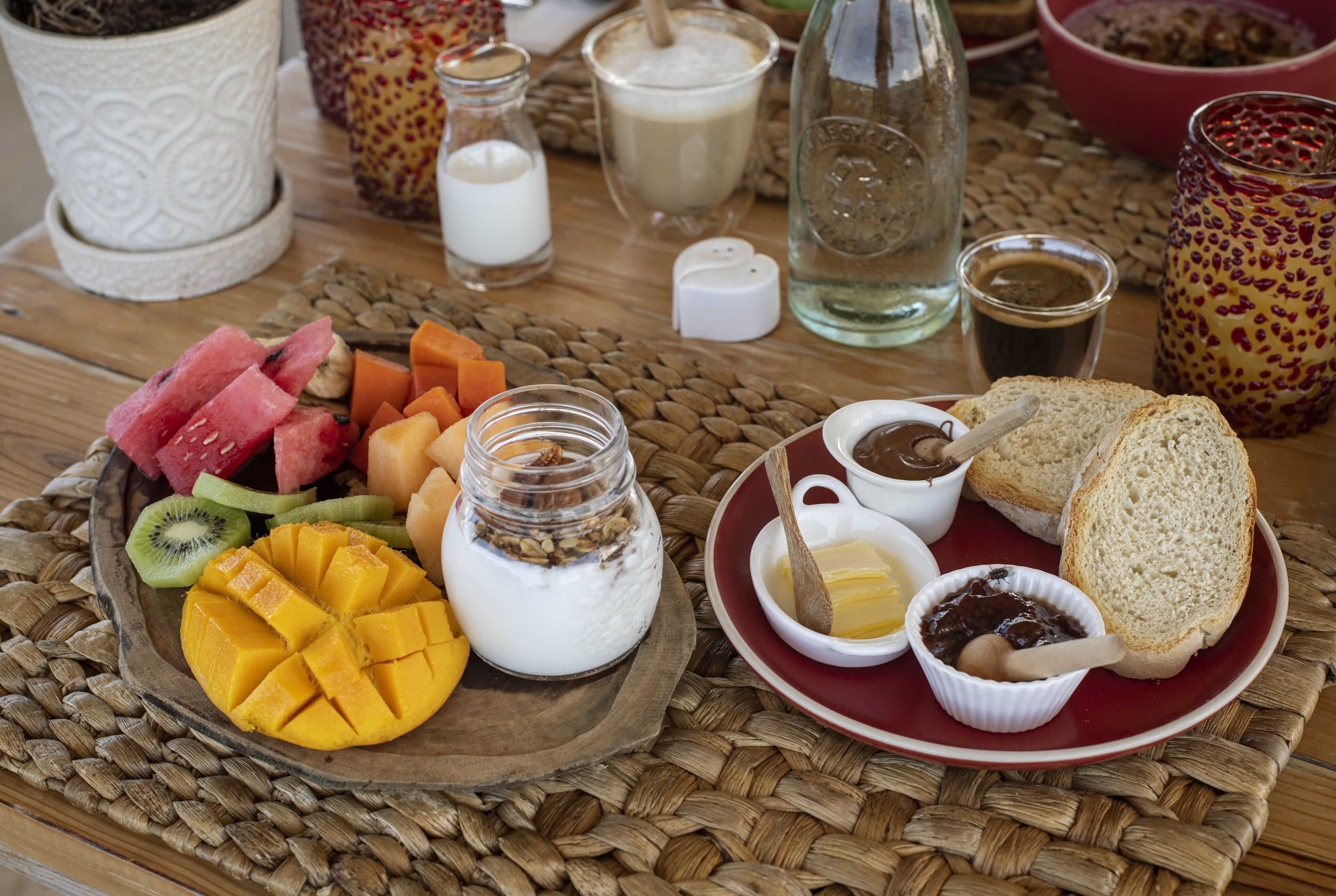 A breakfast spread with fruits, yogurt, bread, and condiments on a woven placemat.