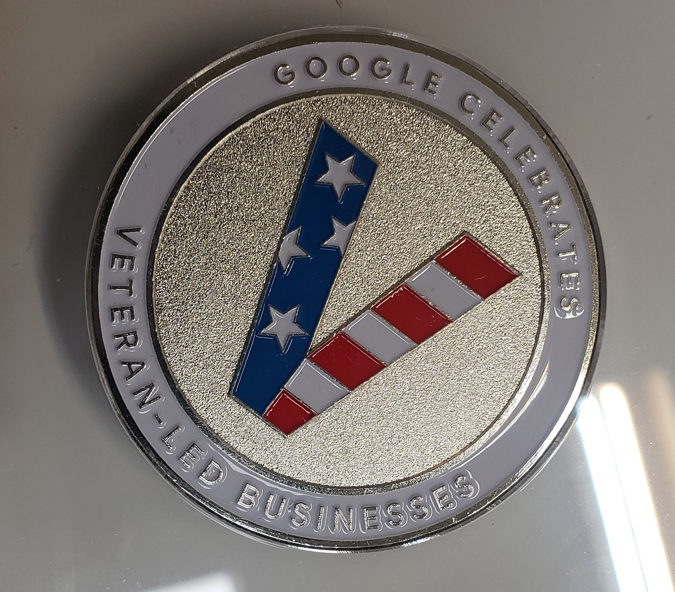 A circular emblem with a textured silver background, featuring a blue and red striped design with stars, and text reading "GOOGLE CELEBRATES VETERAN-LED BUSINESSES".