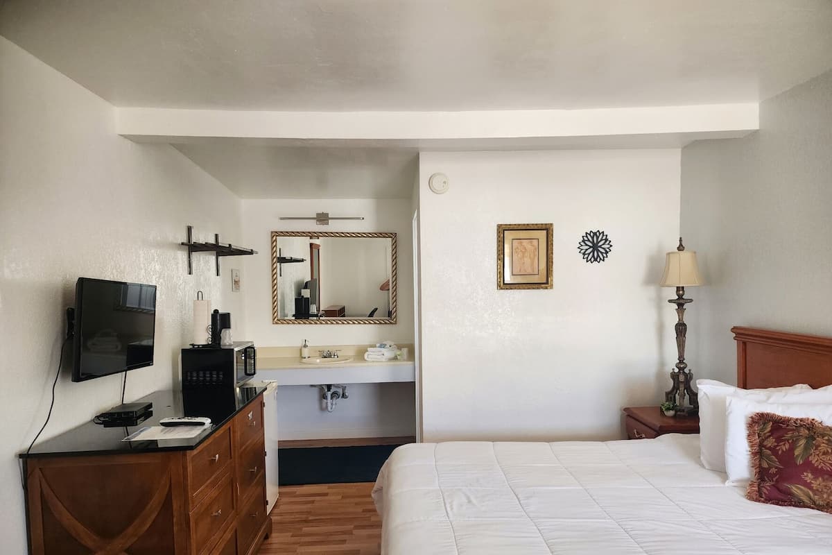 A hotel room with a bed, a desk with a TV, a mirror, a sink, and a bedside table with a lamp.