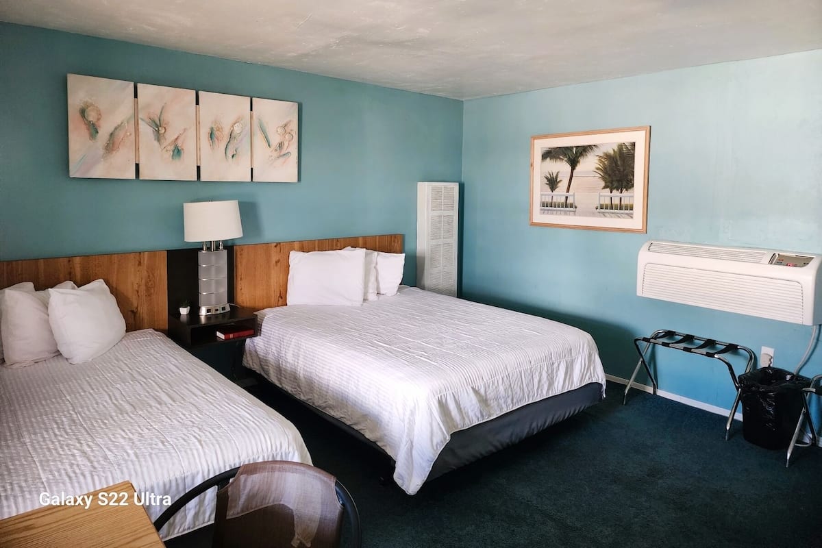 A hotel room with two beds, a nightstand, a lamp, a framed picture, an air conditioner, and a trash bin.