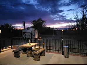 An outdoor seating area with a stone table and benches, a small building with signage, and a metal fence.