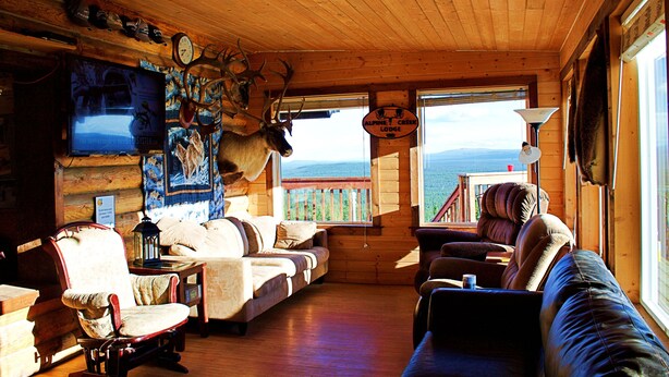 A cozy cabin interior with a wooden ceiling, a large window offering a scenic view, a mounted deer head, and a television on the wall. - Alpine Creek Lodge (Cantwell)