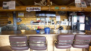A rustic bar area with wooden walls, a stone pillar, and a 'TRIPLE BOB BAR' sign. There are four leather bar stools and a variety of bottles and glasses on the counter. - Alpine Creek Lodge (Cantwell)