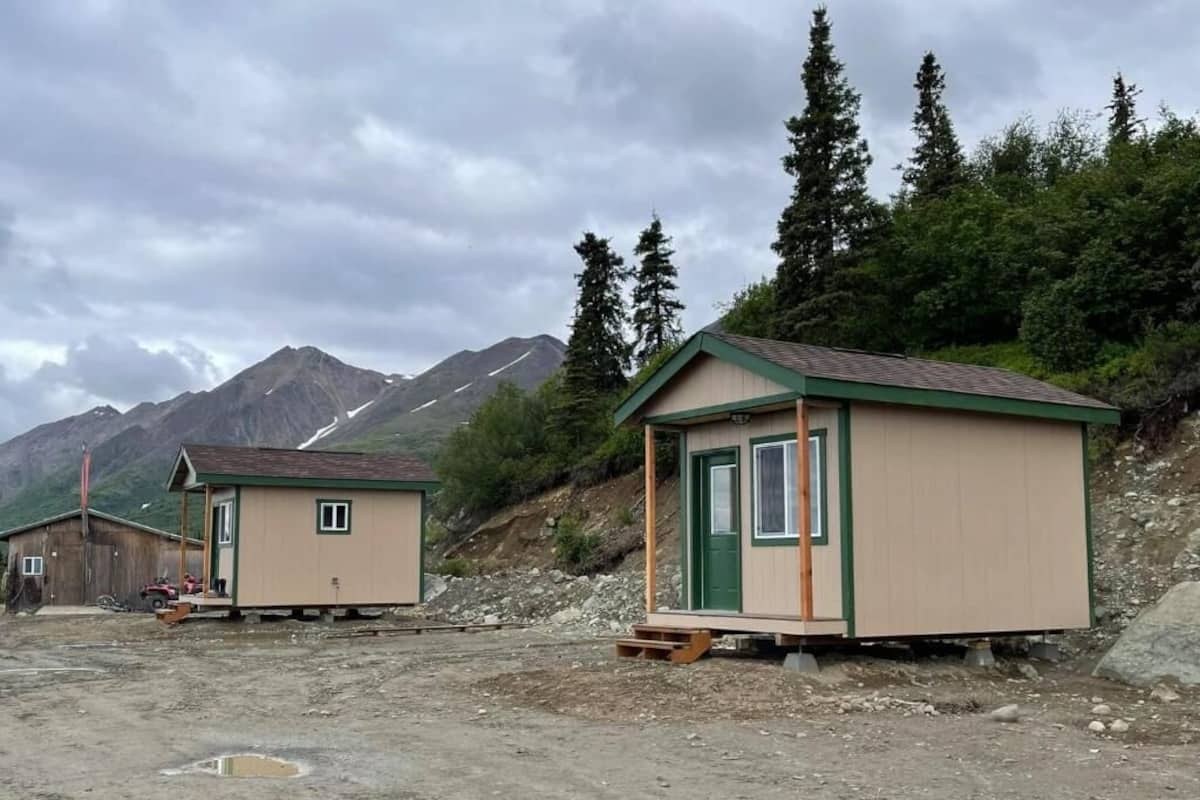 Deux petites cabanes à un seul étage, dotées de boiseries vertes et d’un toit à deux versants, sont construites sur un terrain en terre, avec une chaîne de montagnes en arrière-plan.