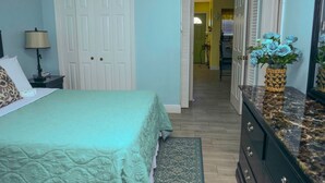A bedroom with a bed, a dresser, a lamp, and a vase of flowers. - Jubilee Gardens Freeport (Freeport)