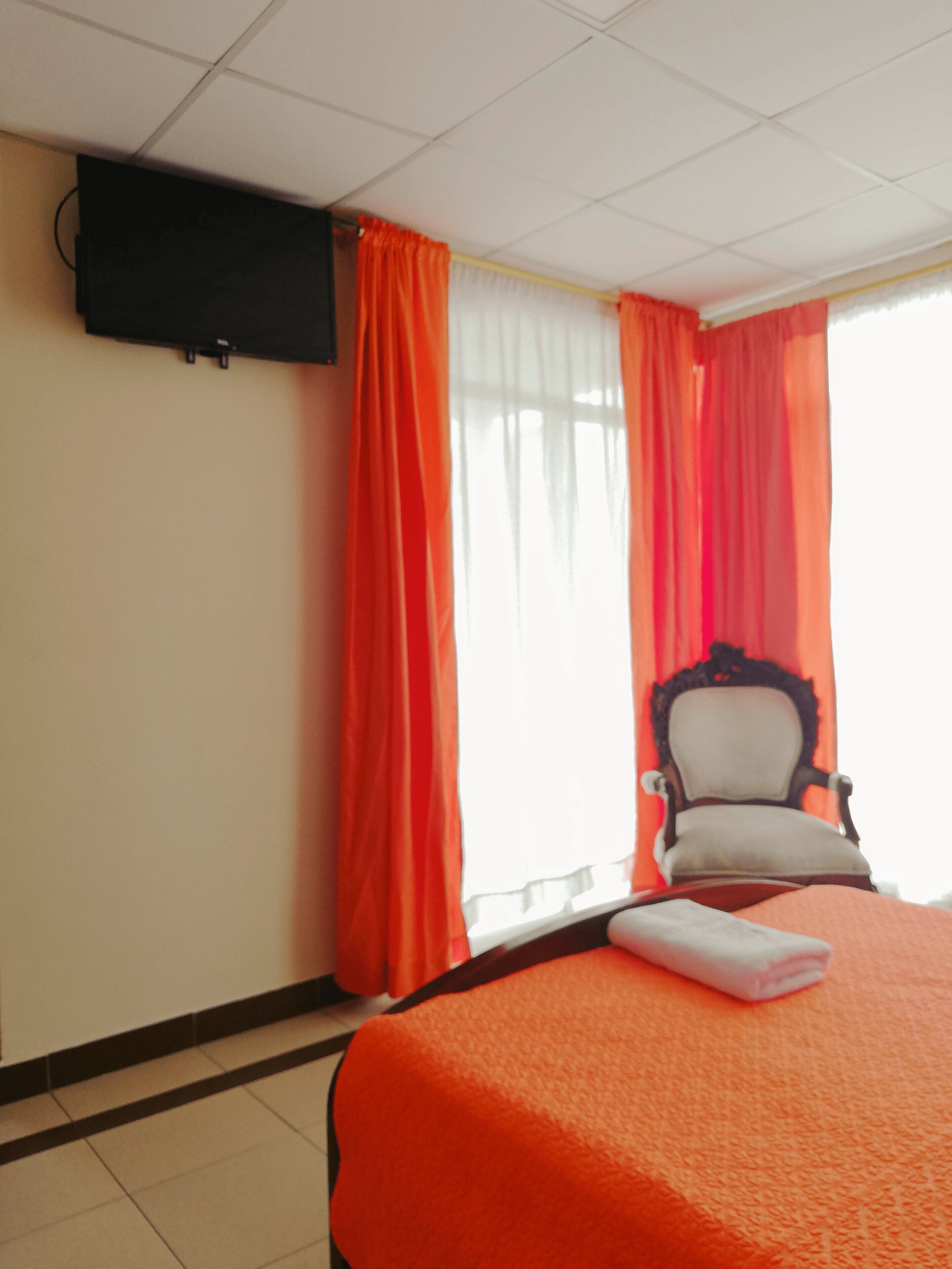 A hotel room with a bed, a chair, a television mounted on the wall, and a window with curtains.