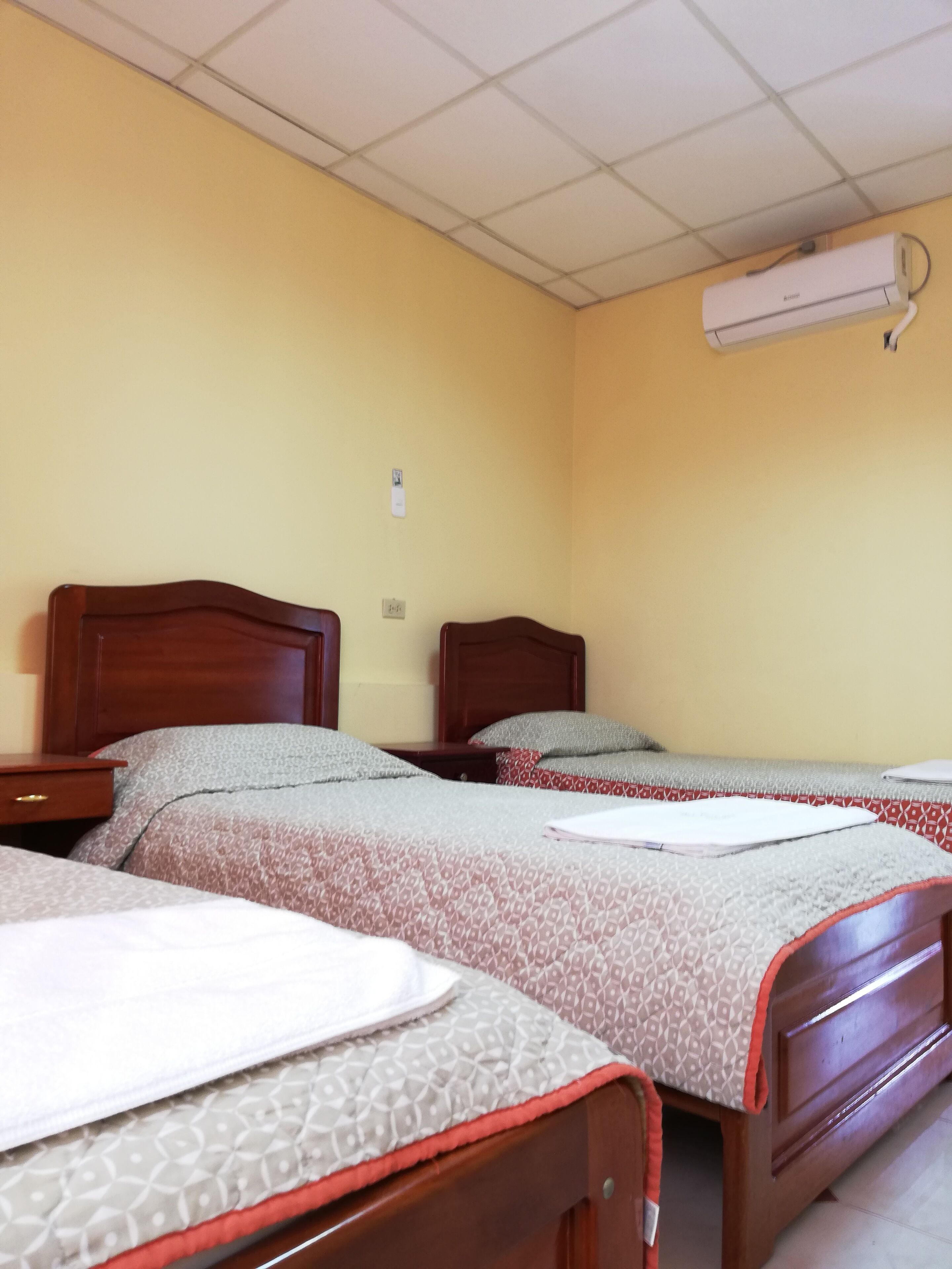 Habitación de hotel con dos camas de madera, cada una con cubrecama y almohadas estampadas. Hay una mesita de noche con una lámpara y un aire acondicionado instalado en la pared.