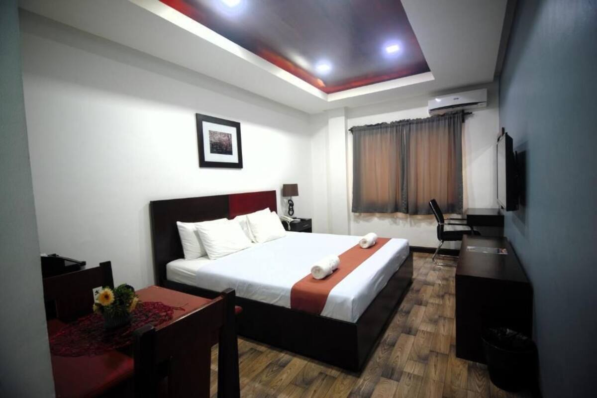 Quarto de hotel com cama, escrivaninha, cadeira, televisão e janela com cortinas.