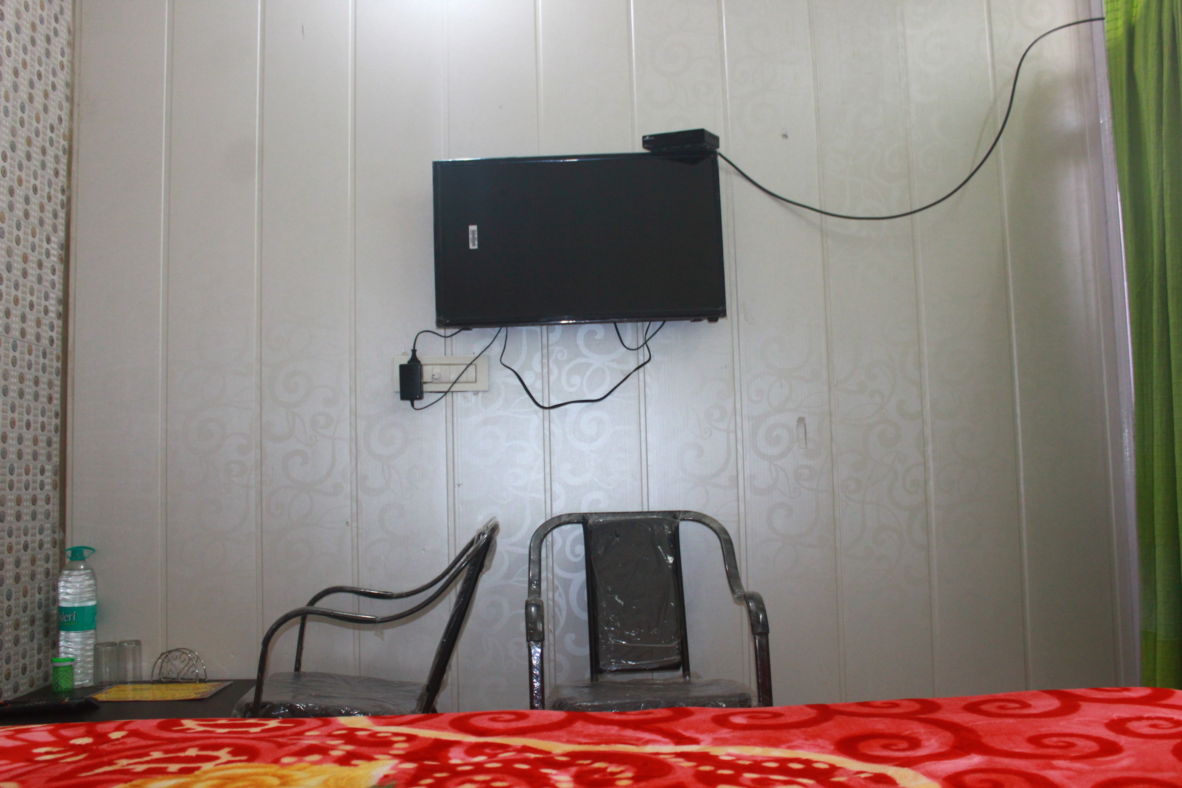 Habitación de hotel con televisión montada en la pared, una silla, una cama con colcha roja estampada y una cortina verde.