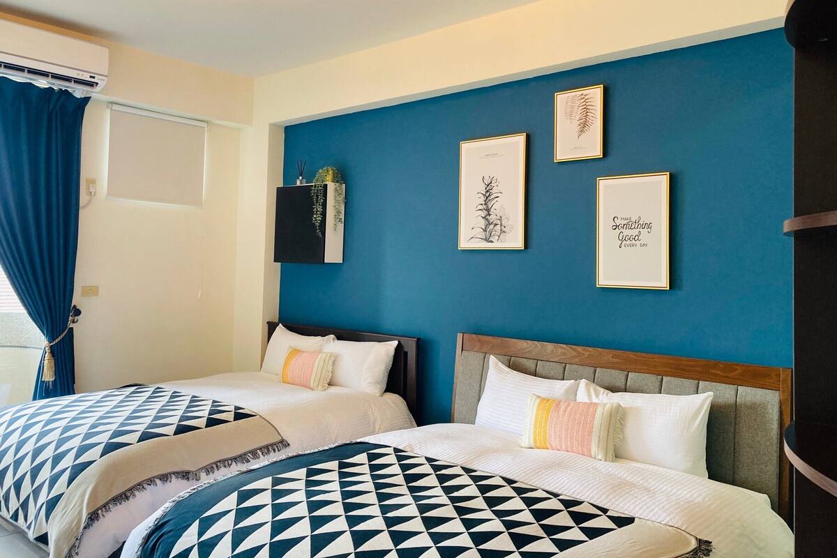 A hotel room with two beds, a blue accent wall, and framed botanical prints.