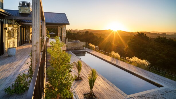 A modern house with a wooden deck, a swimming pool, and a view of the sunset over a hilly landscape. - Te Arai Lodge (Te Arai)