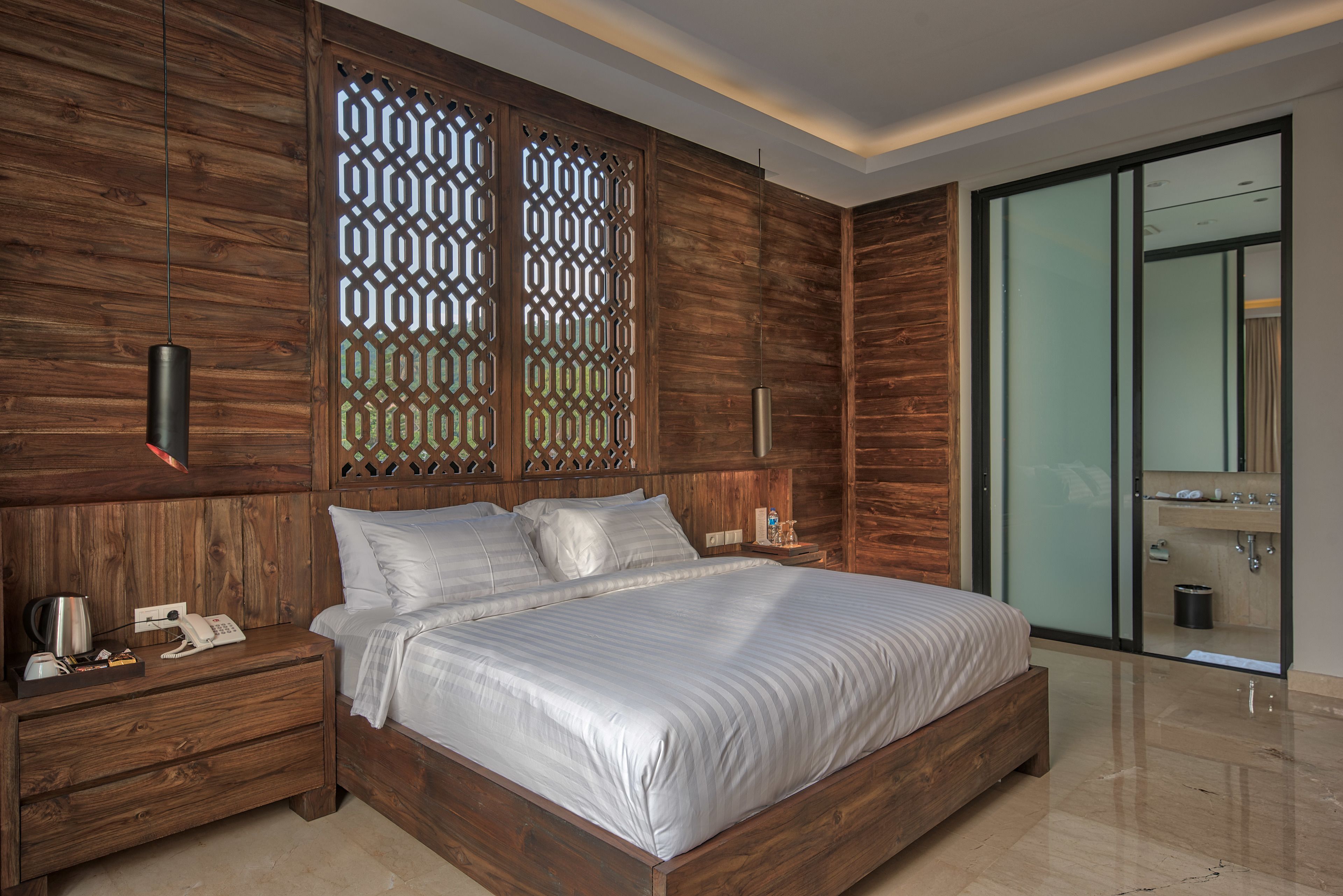 Un dormitorio moderno con cabecera de madera, una cama grande con ropa blanca, una mesita de noche con teléfono y lámpara, y una zona de ducha de vidrio.