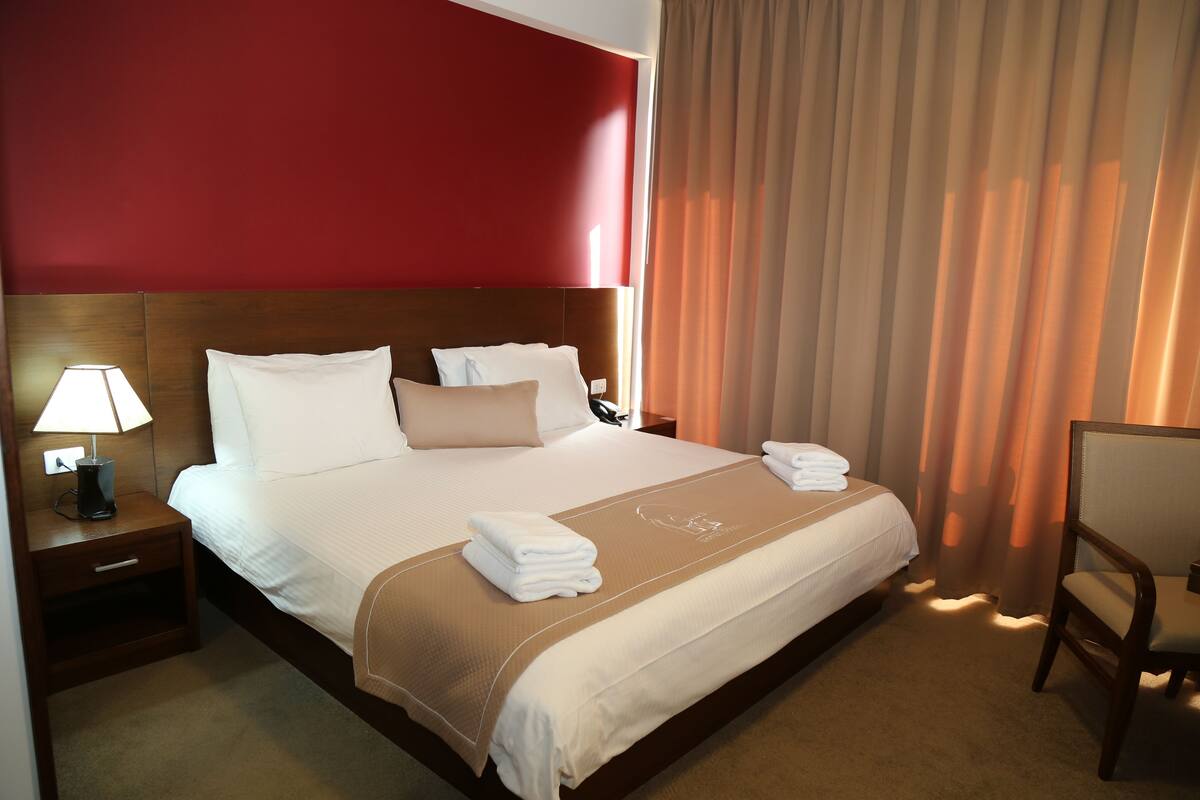 A hotel room with a large bed, a nightstand with a lamp, a chair, and a window with curtains.