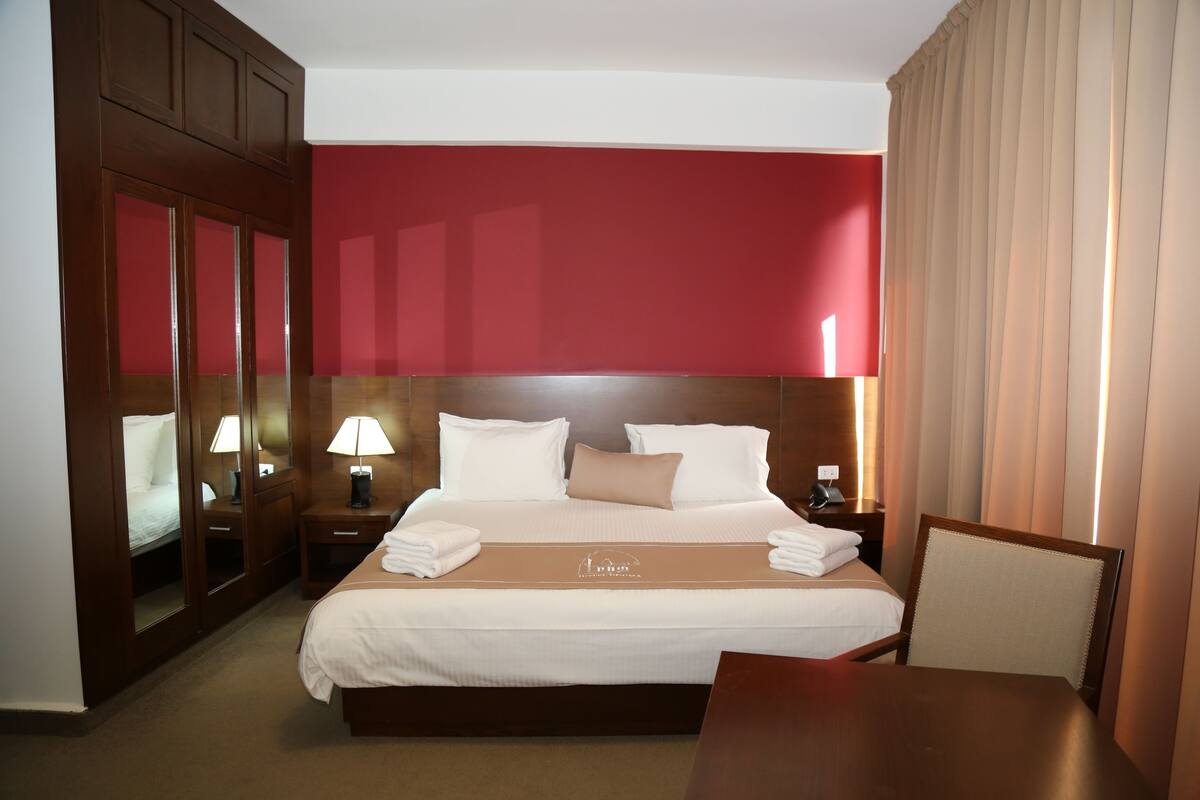 A hotel room with a large bed, a wooden nightstand, a table, and a chair.