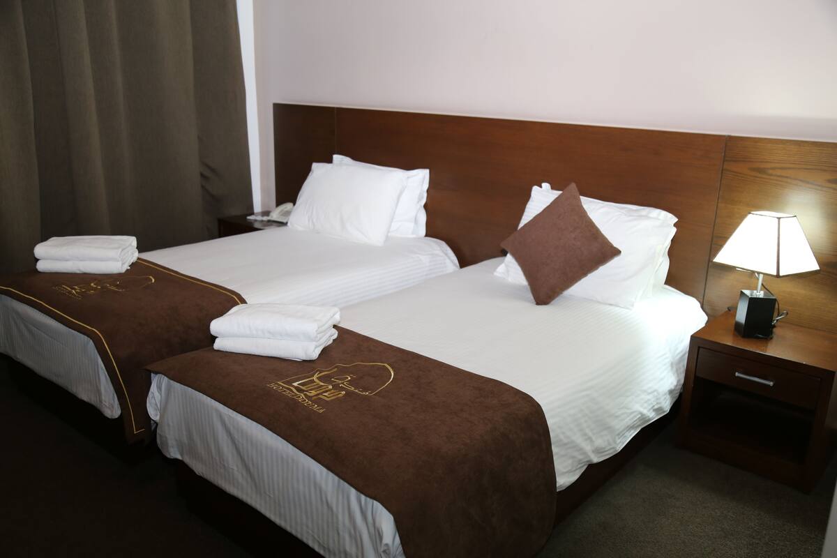 A hotel room with two beds, each with a brown headboard, white linens, and a brown throw blanket. There is a nightstand with a lamp and a drawer.