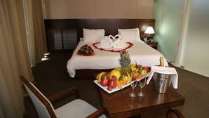 Ein Hotelzimmer mit einem ordentlich bezogenen Bett, einer Obstkorb, einer Champagnerflasche und einem Tisch mit einem Stuhl.