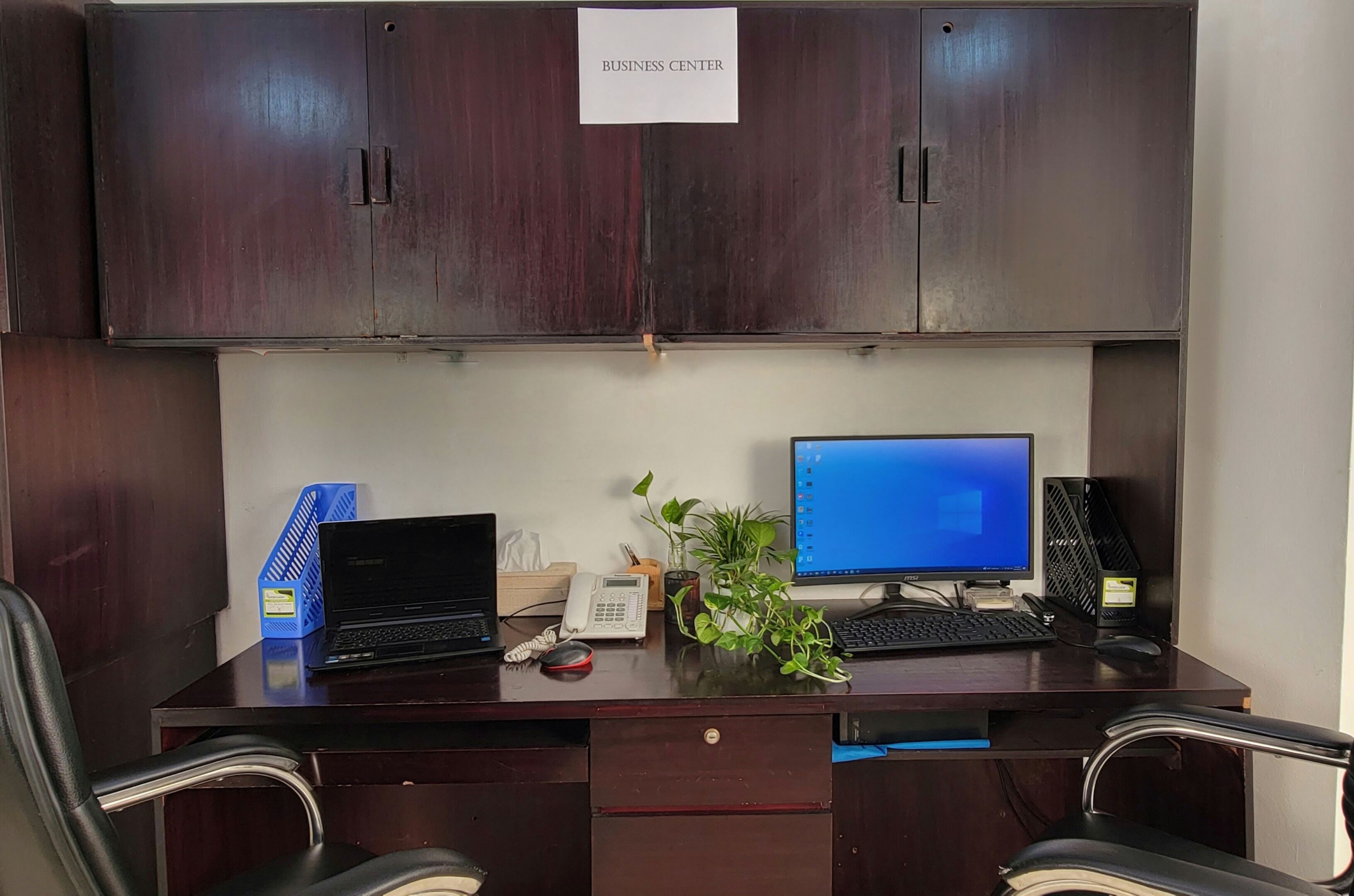 A business center with a desk, two computers, a phone, a plant, and a sign indicating the business center.