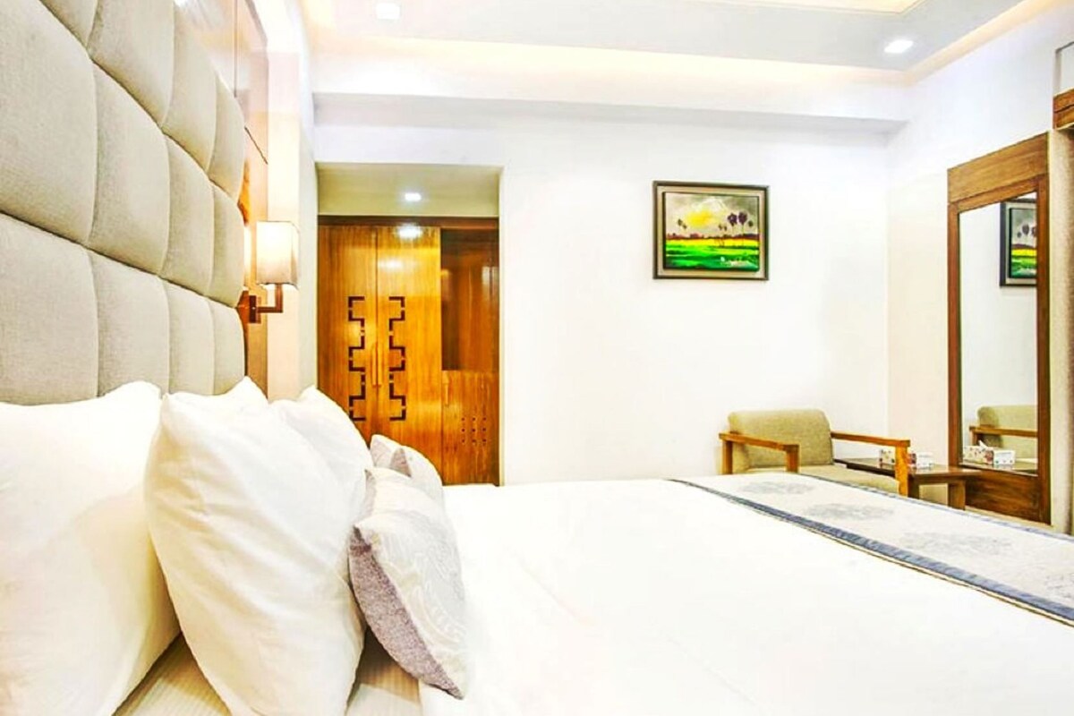 Habitación de hotel con una cama grande, un armario de madera, un cuadro en la pared y una pequeña mesa con una silla.