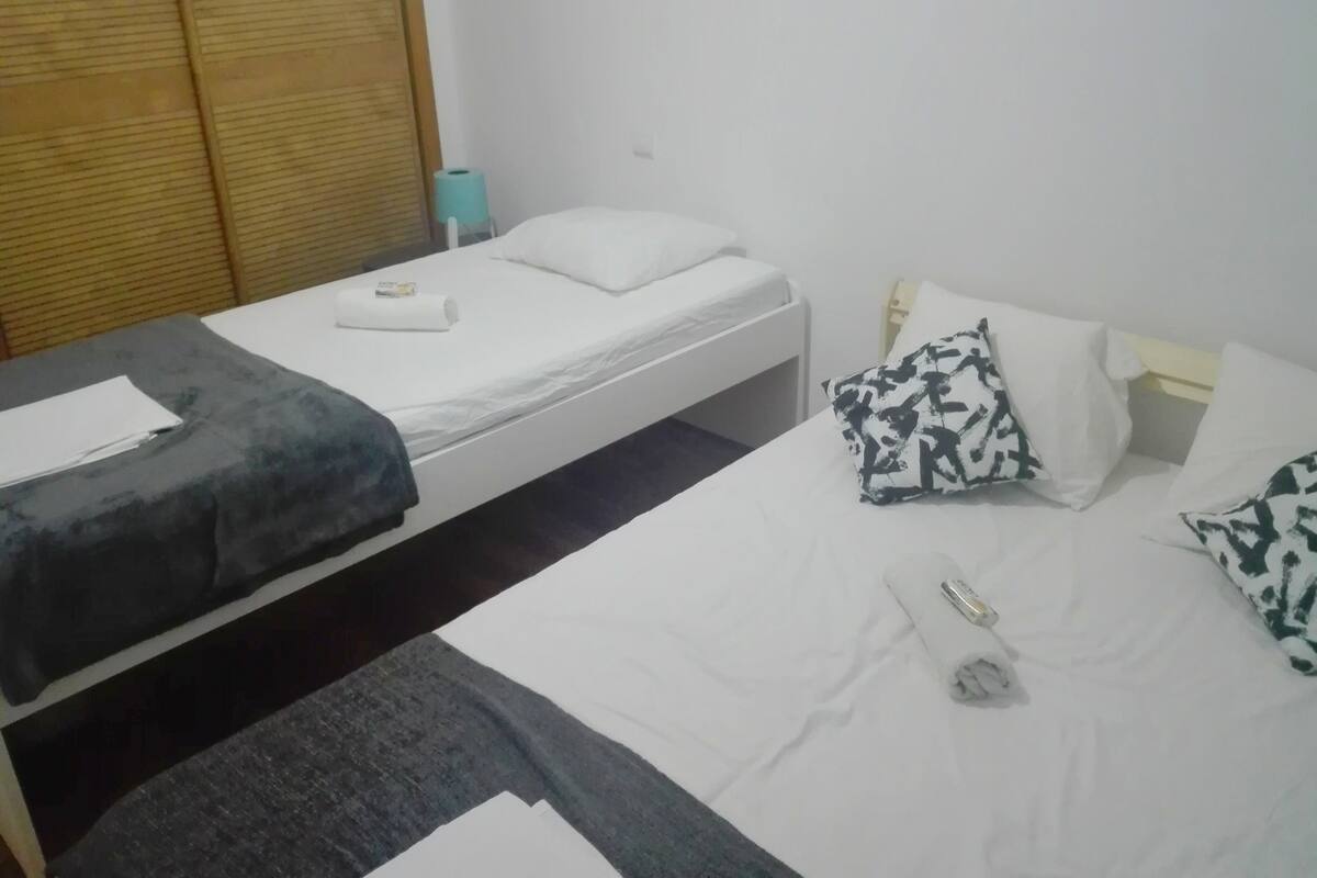 Quarto duplo com duas camas de solteiro, um guarda-roupa de madeira, uma mesinha e uma mesadita.