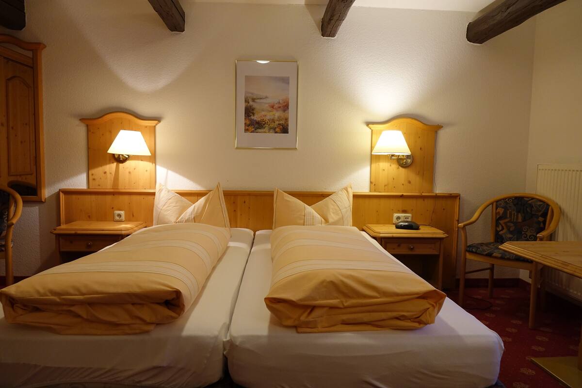 A hotel room with two beds, wooden headboards, bedside tables with lamps, a framed picture on the wall, and a chair.