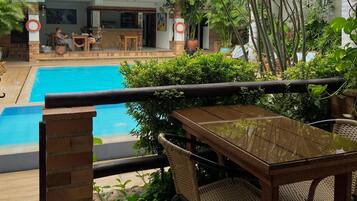 Zona junto a la piscina con mesa y sillas, rodeada de abundante vegetación y un edificio al fondo.