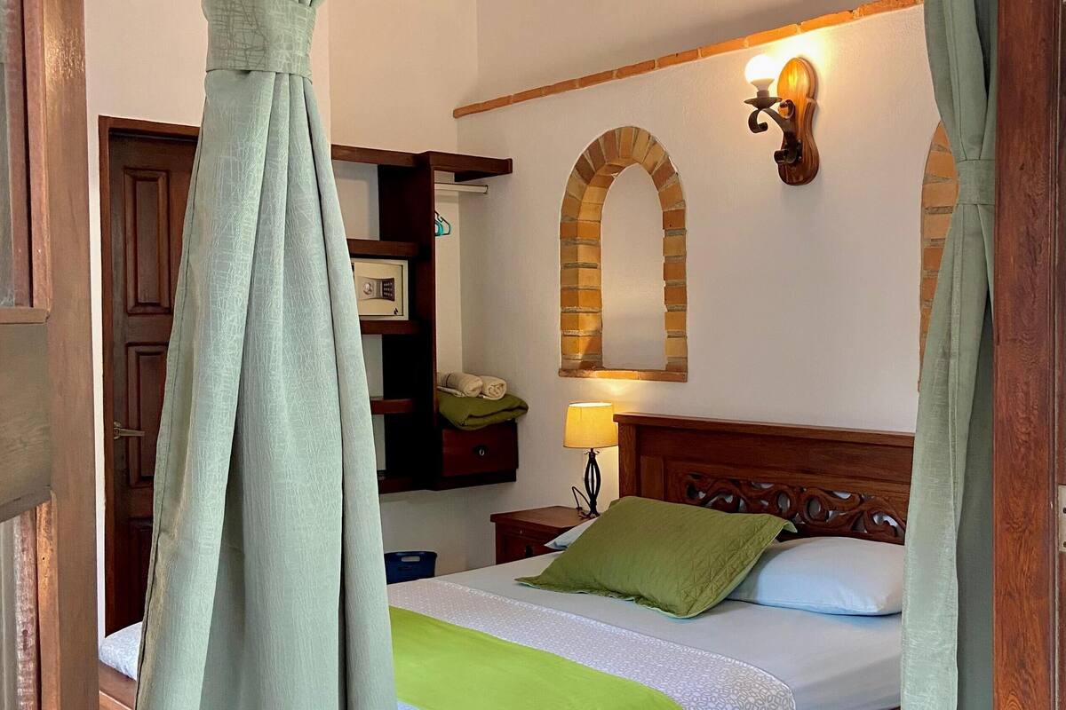 A bedroom with a wooden bed, green curtains, a wall-mounted lamp, and a brick-patterned shelf.