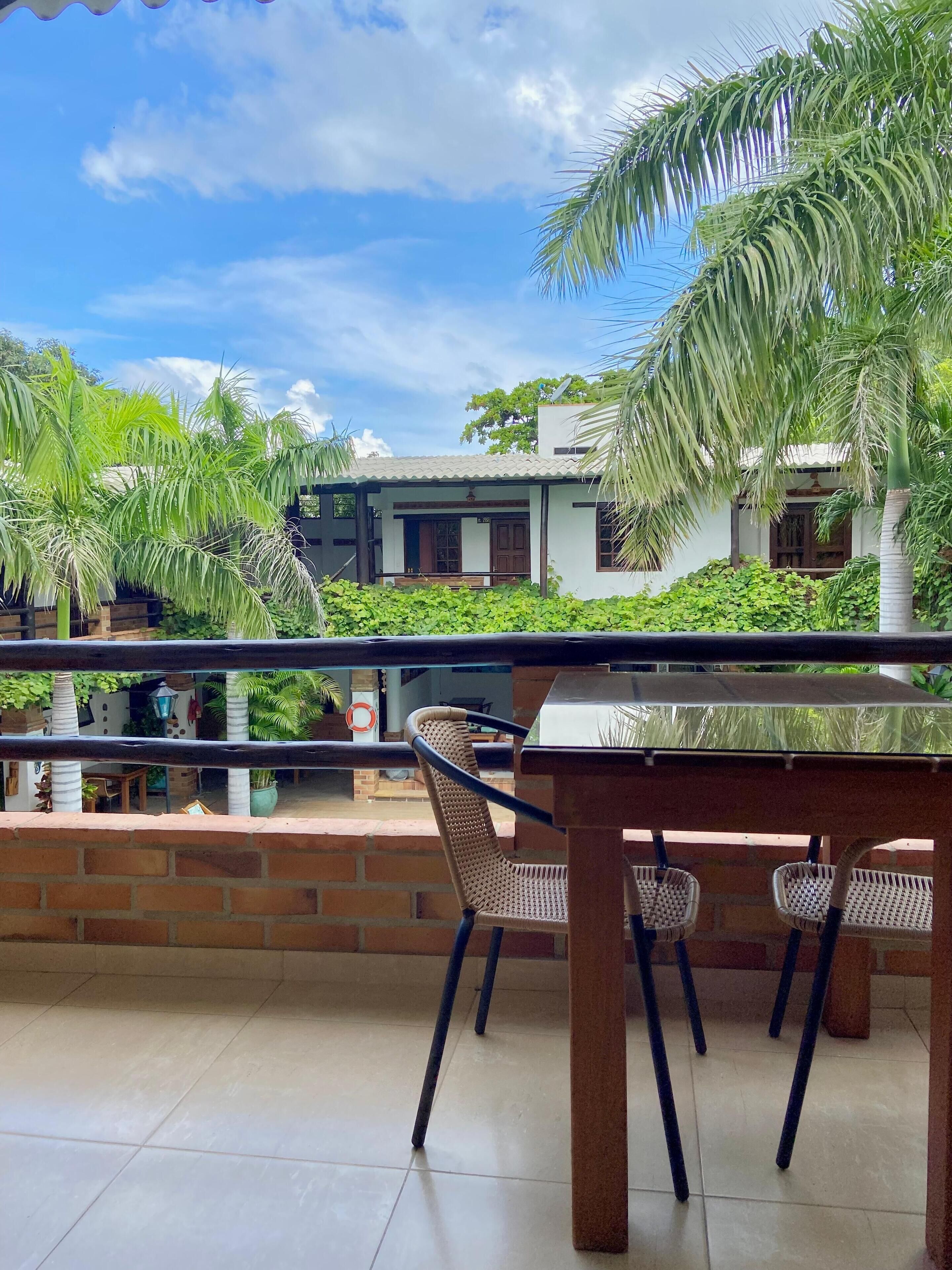 Ein Balkon mit Tisch und Stühlen, der auf ein Gebäude mit Ziegeldach blickt und von Grünpflanzen umgeben ist.