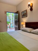 A hotel room with a bed, a nightstand with a lamp, a wooden headboard, a framed picture on the wall, and an open door leading to a balcony with outdoor furniture.