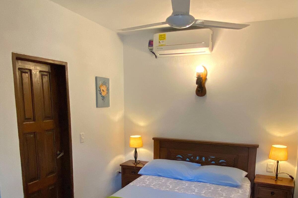 A bedroom with a wooden bed, two bedside tables with lamps, a ceiling fan, and a wall-mounted air conditioner.