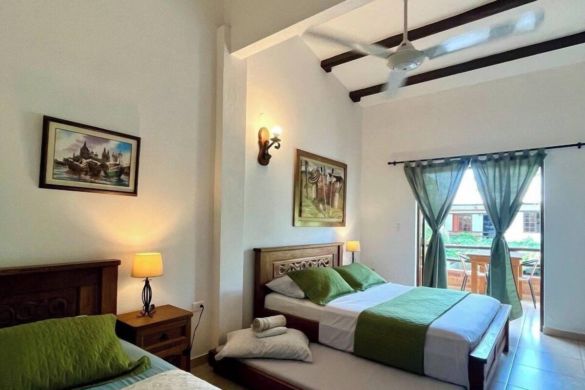 A hotel room with two beds, a ceiling fan, a painting on the wall, and a view through a window with curtains.
