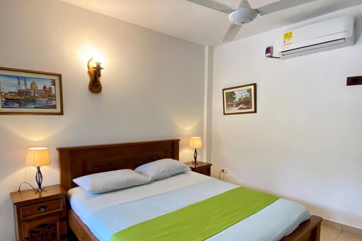 A neatly made bed with a green blanket, wooden headboard, two bedside tables with lamps, a ceiling fan, and wall-mounted air conditioning.