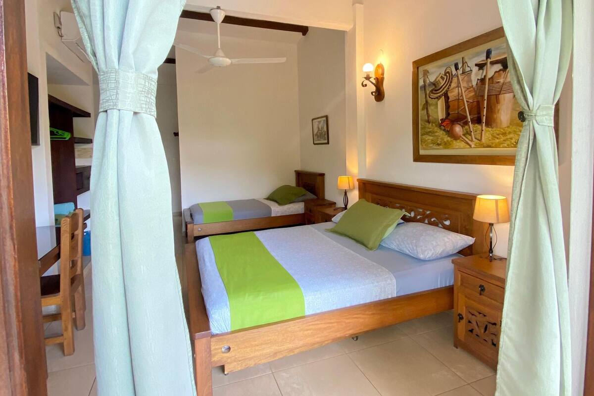 A bedroom with a wooden bed, two bedside tables, a nightstand, a ceiling fan, a painting on the wall, and a dining area with a chair and table.