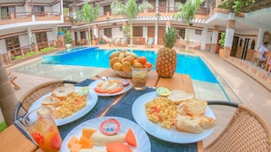 Desayuno con platos de comida, una piña y un vaso de zumo de naranja junto a una piscina.