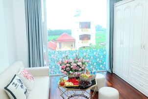 Quarto com sofá branco, mesa com cesta de frutas, vaso de flores e vista para um edifício.