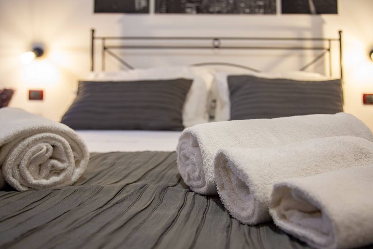 Un lit bien fait, recouvert d’une couverture grise et agrémenté de deux serviettes blanches roulées.