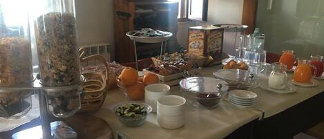 Una mesa de buffet con diversos alimentos y recipientes, incluyendo tazones de frutas, un contenedor de cereales y varias jarras de bebidas.