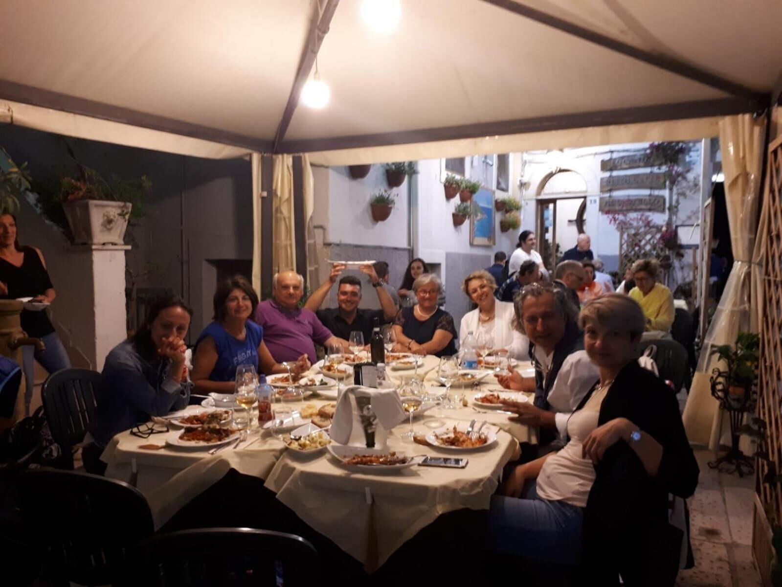 A group of people dining at an outdoor table with white tablecloths, surrounded by potted plants and hanging decorations.