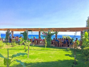 An outdoor seating area with tables and chairs under a wooden pergola, overlooking a body of water. - Sahin Motel & Restaurant (Bodrum)