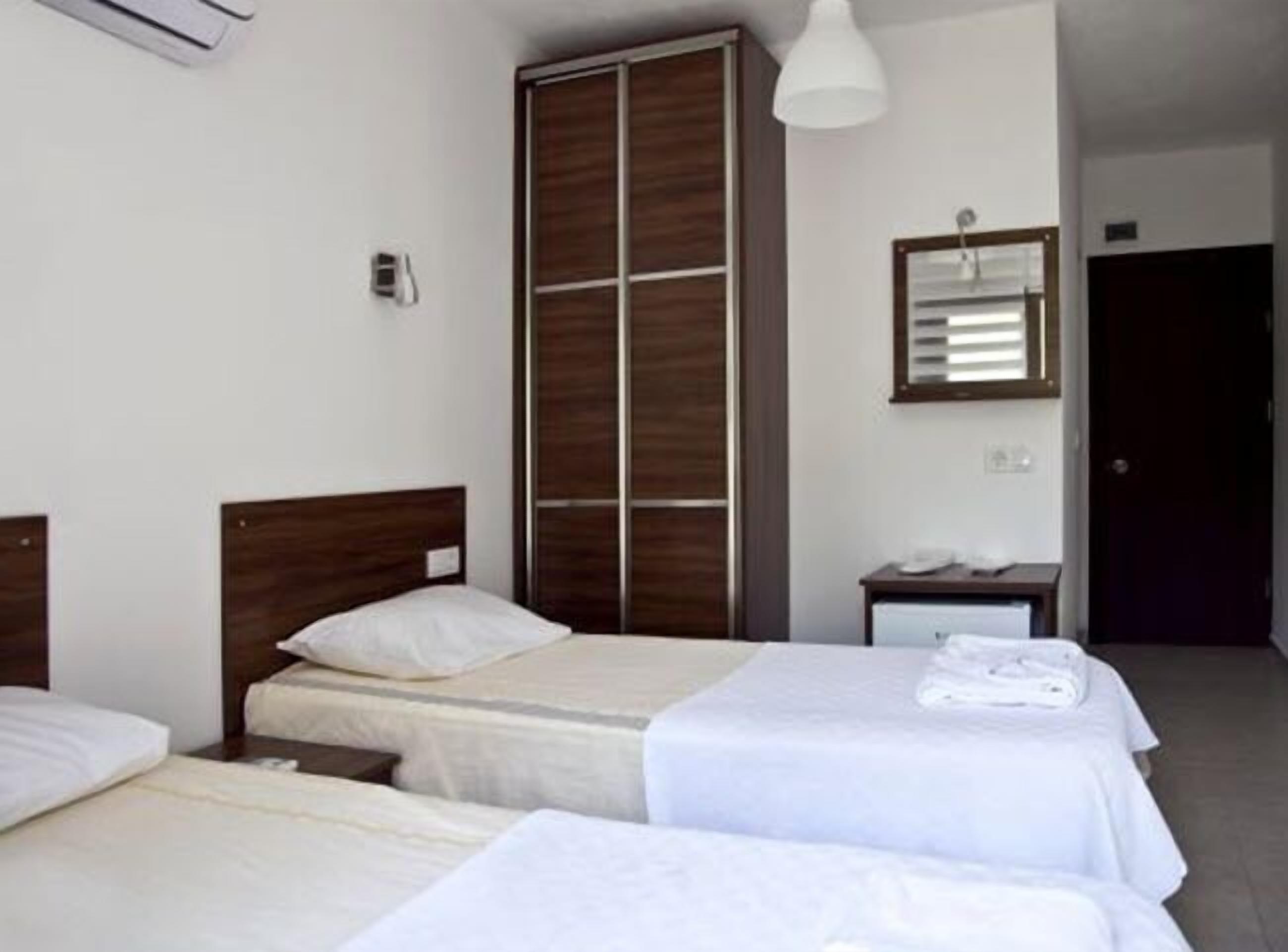 Quarto de hotel com duas camas de solteiro, um guarda-roupa, uma mesa de cabeceira com abajur e um espelho na parede.