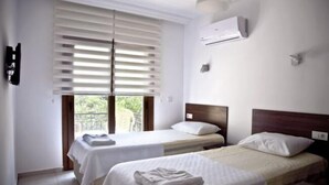 A hotel room with two beds, a window with blinds, an air conditioning unit, and a wall-mounted TV. - Sahin Motel & Restaurant (Bodrum)
