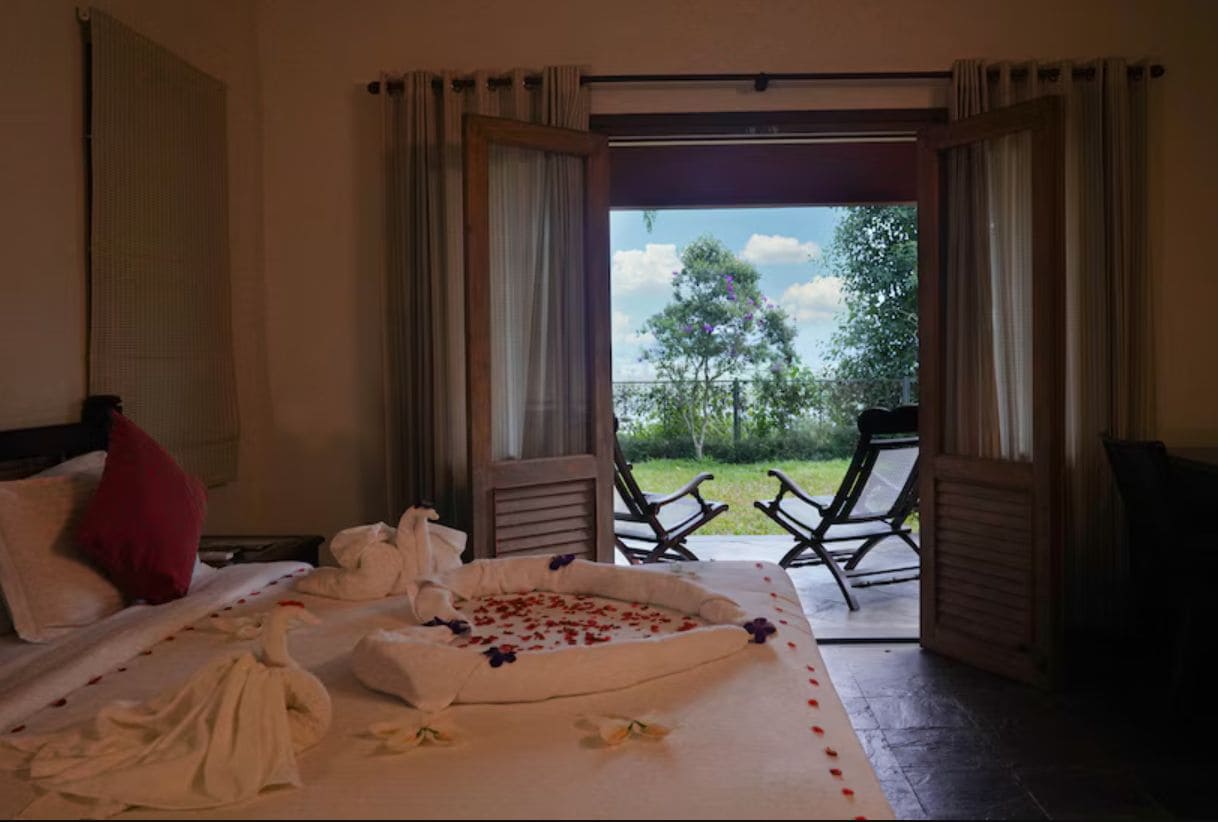 Habitación de hotel con una cama, una almohada roja, una toalla y un arreglo floral. Cuenta con un balcón con sillas y vistas a árboles.
