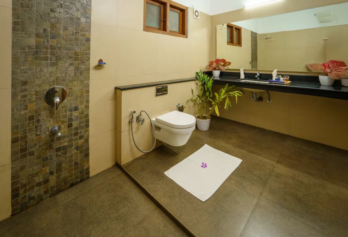 A bathroom with a tiled shower wall, a toilet, a sink with a mirror above, and a potted plant on the counter.