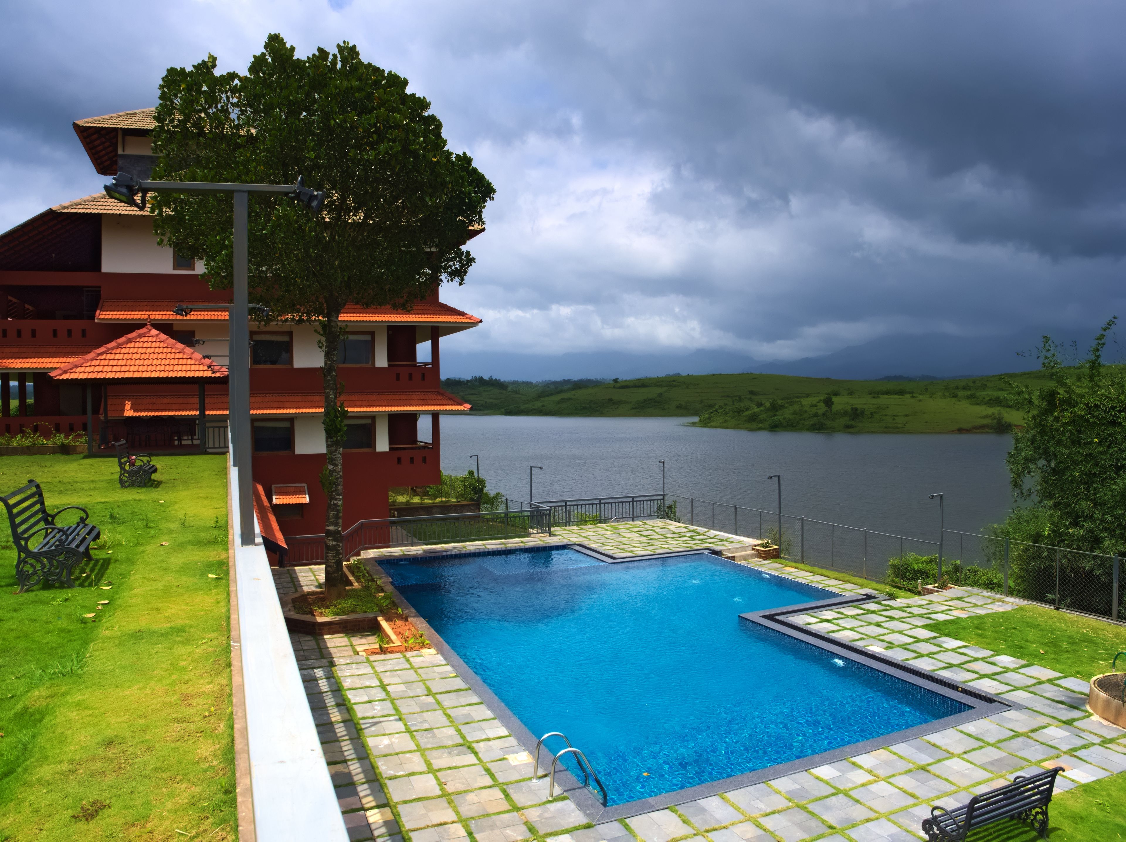 A swimming pool with a tiled deck, a bench, and a view of a lake and a building with a red roof.