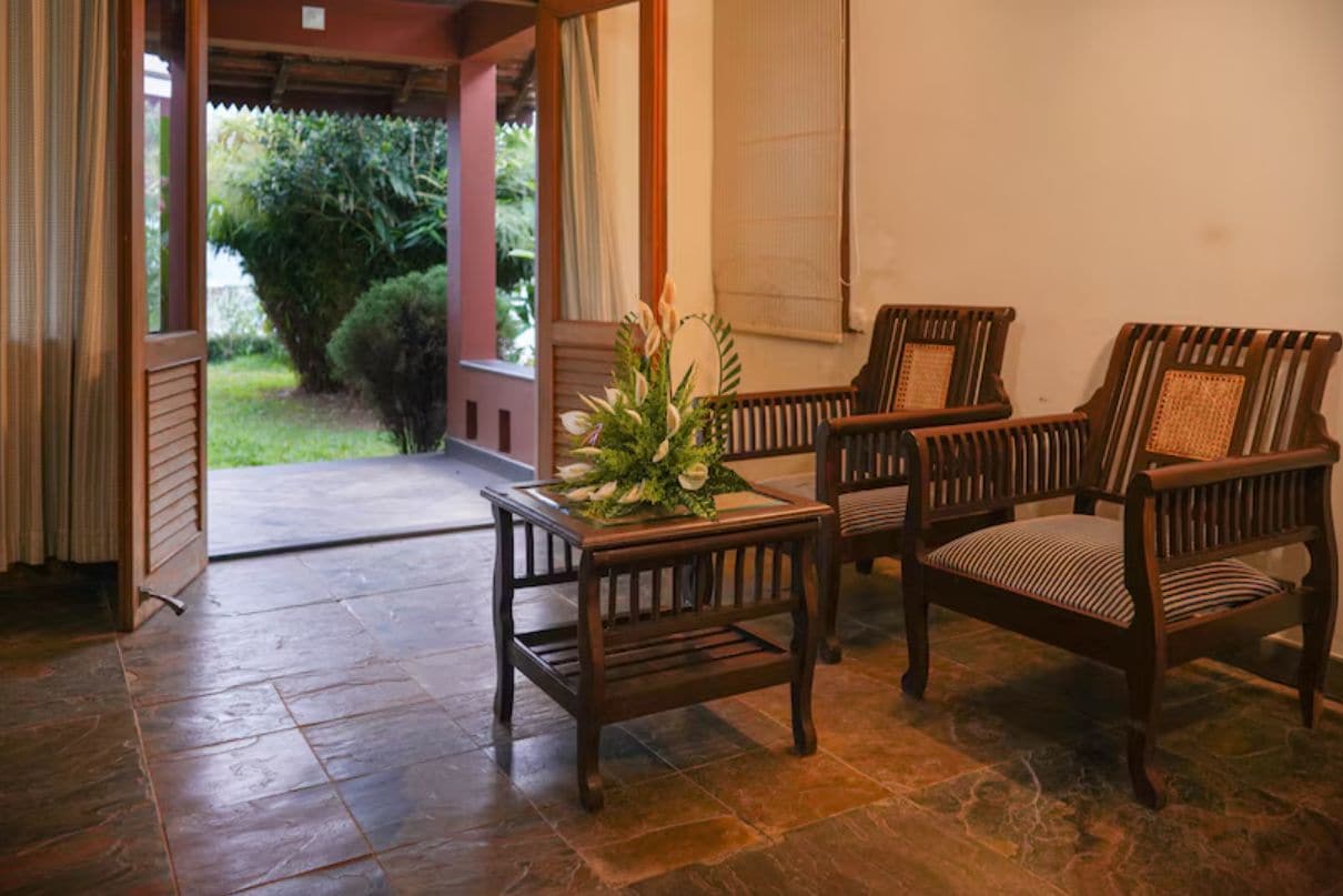 Una habitación con una mesa de madera y dos sillas, una planta en maceta y vistas a un jardín a través de una puerta abierta.