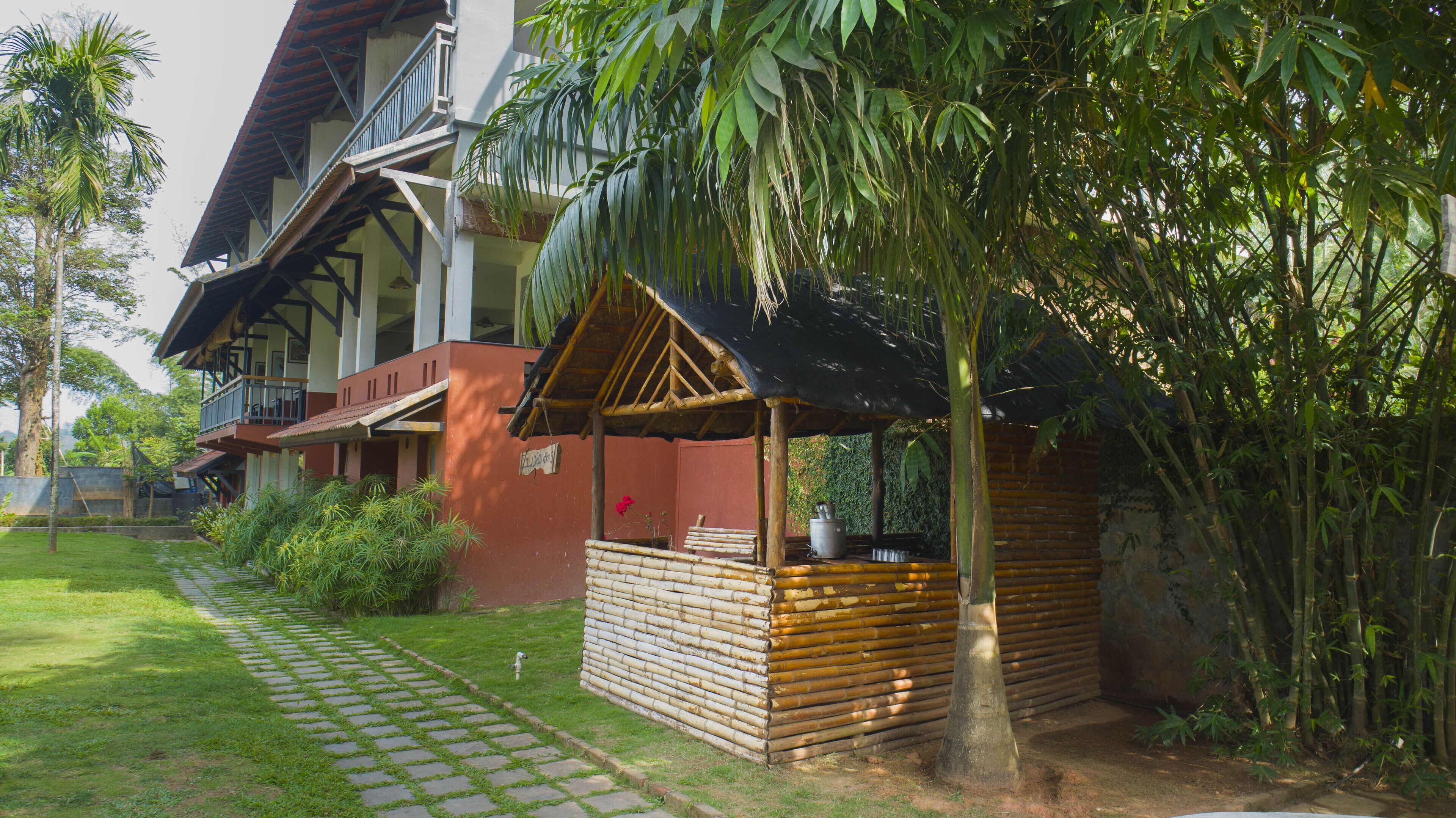 A traditional wooden structure with a thatched roof, surrounded by lush greenery and a paved pathway.