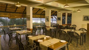 A restaurant with multiple tables and chairs, large windows offering a view of the outdoors, and hanging pendant lights. - Vistara Resort (Sulthan Bathery)