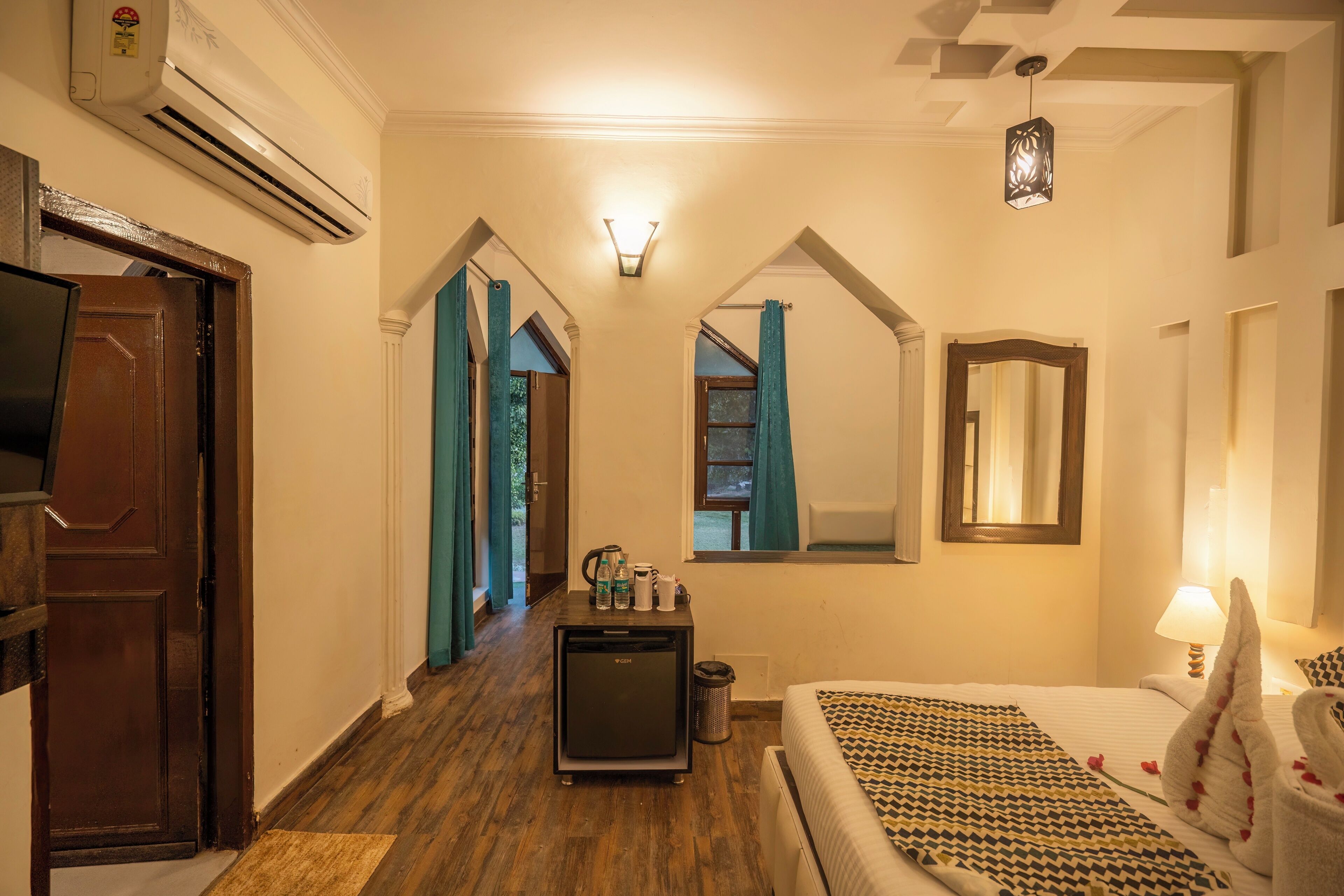 A hotel room with a bed, a desk with a refrigerator, a mirror, and a hallway with a door and curtains.